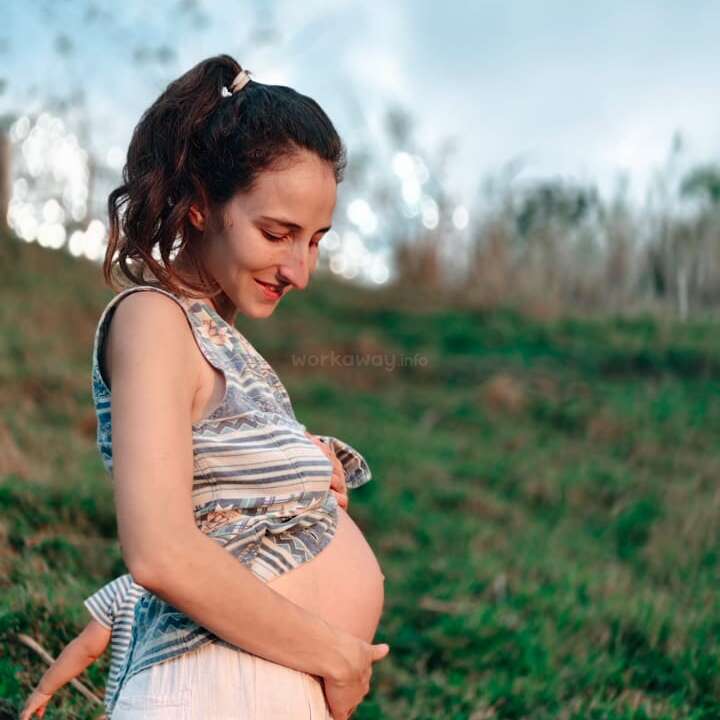 Uma mulher está do lado de fora, em uma colina gramada, sorrindo e segurando gentilmente sua barriga de grávida, vestindo uma blusa listrada sem mangas e uma saia branca. Ambiente tranquilo e natural.