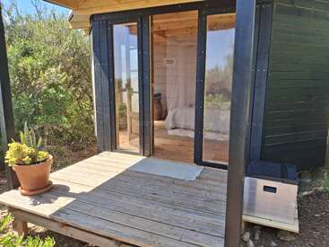 A small wooden cabin with a glass door opens to a cozy bedroom. A potted plant and storage box decorate the sunlit front porch surrounded by greenery.