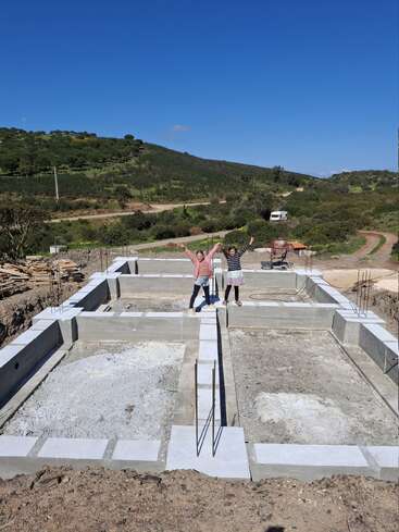 Zwei Menschen stehen voller Vorfreude auf dem Fundament eines neuen Gebäudes, umgeben von malerischen grünen Hügeln, strahlend blauem Himmel und Baumaterialien an einem sonnigen Tag.