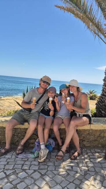 Eine lächelnde vierköpfige Familie sitzt auf einer Bank am Meer und genießt unter strahlend blauem Himmel Eiswaffeln, umgeben von Palmen und Glück.