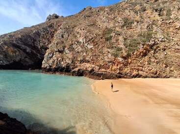 Eine Person steht an einem ruhigen Sandstrand am türkisfarbenen Wasser. Im Hintergrund erheben sich steile Felsklippen unter einem strahlend blauen Himmel. Friedliche Atmosphäre.
