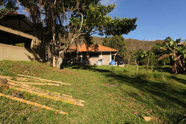 Una casa rural con tejado de tejas rojas se encuentra entre hierba verde, tallos de bambú, árboles y montañas bajo un cielo azul despejado en un día soleado.