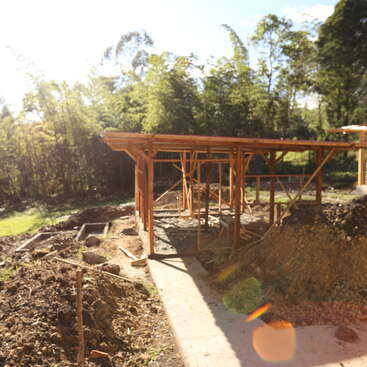 A wooden framework of a building under construction stands on a sunny plot, surrounded by dirt piles, green grass, trees, and a bright lens flare.