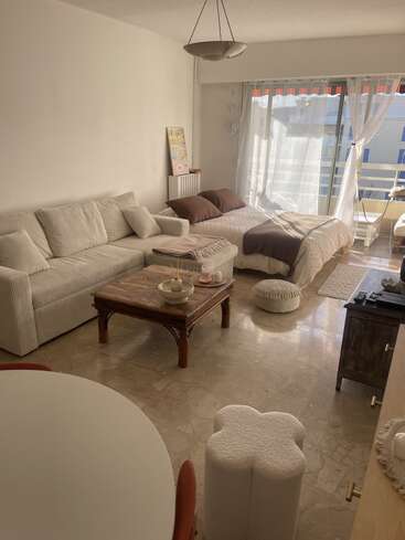 Ce salon chaleureux est aménagé avec un canapé beige, une table basse en bois, un lit, des poufs, une table ronde, et bénéficie d'une lumière naturelle provenant de grandes fenêtres, ainsi que de rideaux vaporeux qui créent une ambiance douce.