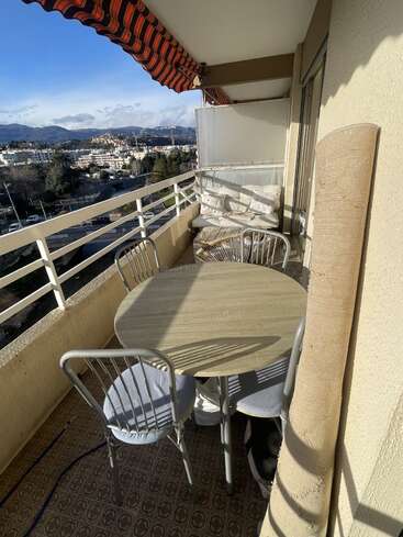 Un balcon ensoleillé avec une table ronde et trois chaises, un store rayé, un banc rembourré et un tapis enroulé ; il donne sur un panorama urbain pittoresque avec des collines au loin.