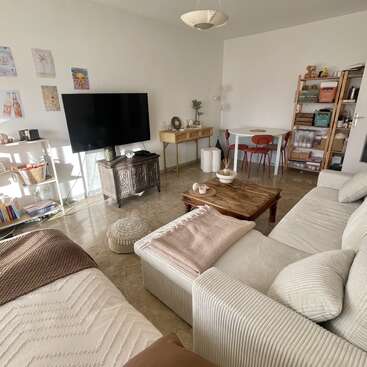 Bright cozy living room with a large TV, comfortable sofa, wooden coffee table, small dining area, shelves, light décor, and wall art. Inviting atmosphere.