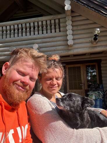 Un homme barbu, une femme souriante et un chien noir profitent du soleil sur un porche devant une cabane rustique en rondins, dégageant chaleur, confort et bonheur.
