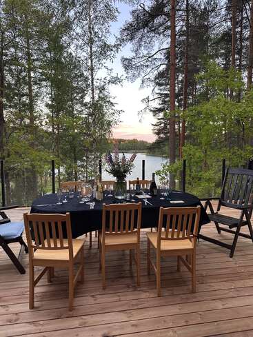 Ein wunderschön gedeckter Esstisch im Freien auf einer Holzterrasse mit Blick auf einen ruhigen See, umgeben von hohen grünen Bäumen unter einem friedlichen Abendhimmel. Friedliche Atmosphäre.