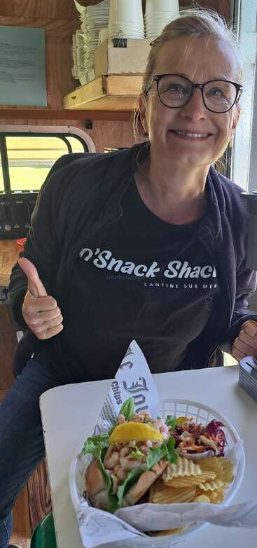 Une femme souriante portant des lunettes et un tee-shirt "Snack Shack" lève le pouce, assise à côté d'un panier de nourriture contenant des chips, de la salade et un sandwich.