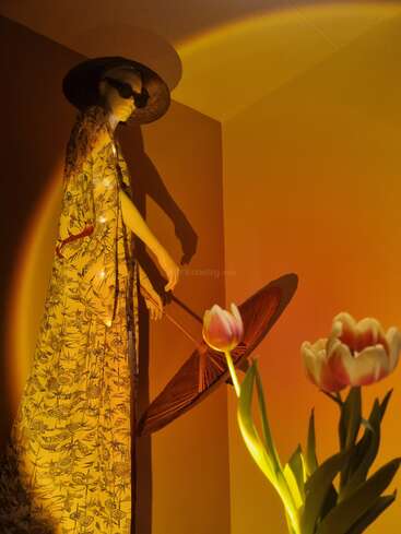 The image features a mannequin wearing a yellow floral dress, holding an umbrella, and standing beside a vase of tulips in a dimly lit room with a warm, golden glow.