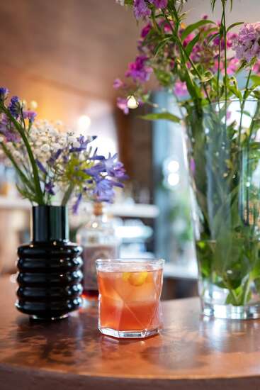 L'image représente un verre de liquide orange posé sur une table en bois, entouré de vases de fleurs, avec un arrière-plan flou suggérant un restaurant ou un bar.