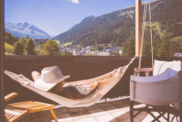 Una persona con un sombrero blanco descansa en una hamaca en un balcón soleado, con vistas a un precioso pueblo de montaña rodeado de frondosos bosques verdes y un cielo azul.