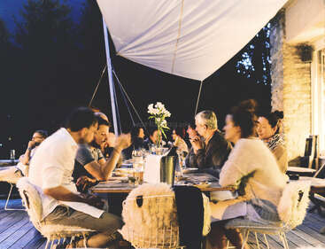 Un grupo de personas disfruta de una cena al aire libre bajo un toldo por la noche. La cálida iluminación crea un ambiente acogedor y festivo, en el que no faltan las risas y la conversación.