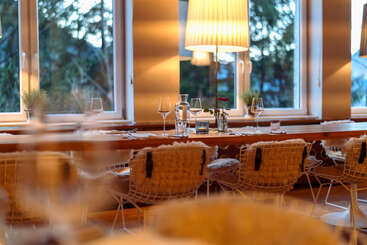 El interior del restaurante, acogedor y con una iluminación cálida, cuenta con una larga mesa de madera, sillas modernas con fundas mullidas, una elegante cristalería y un centro de mesa con una sola rosa roja junto a los grandes ventanales.