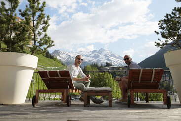 Dos hombres descansan al aire libre en unas tumbonas de madera, rodeados de grandes árboles en macetas, disfrutando de un día soleado con unas impresionantes vistas de las montañas nevadas a sus espaldas.