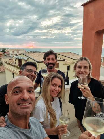 The image shows five people on a balcony overlooking the sea, smiling and holding wine glasses, with a bottle of wine and a plate of food nearby.