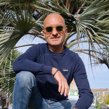 A bald man in sunglasses crouches on grass beneath a palm tree, wearing a navy sweater, jeans, sneakers, and a smartwatch, smiling confidently at the camera.