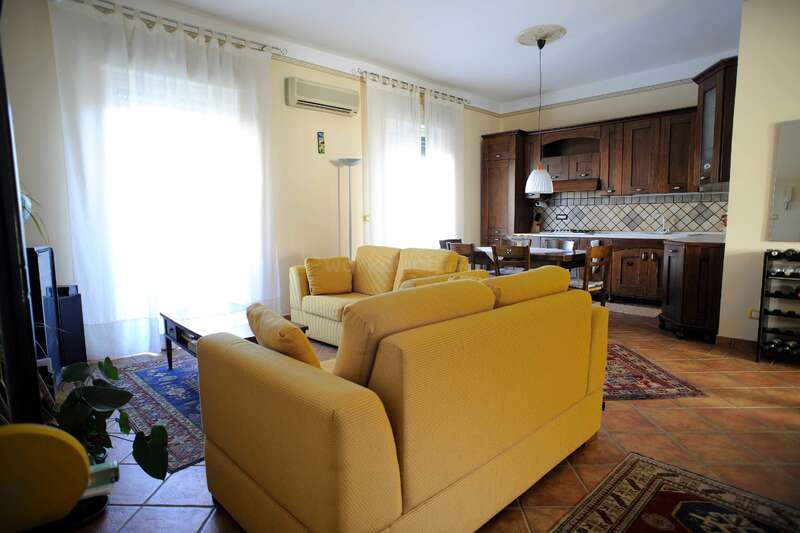 The image depicts a living room with yellow couches, a blue rug, and a TV, surrounded by windows with white curtains, creating a bright and airy atmosphere.