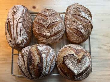 Cinco pães artesanais descansam em uma grade de resfriamento. Cada pão apresenta desenhos exclusivos e intrincados, incluindo folhas, padrões geométricos e um coração.