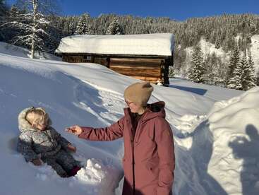 Uma mulher com casaco rosa e chapéu bege ajuda uma criança sentada na neve profunda, ao lado de uma cabana de madeira cercada por árvores nevadas e sol brilhante de inverno.