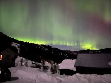 A imagem mostra uma paisagem de neve à noite com casas, árvores e montanhas. O céu é iluminado por impressionantes luzes do norte verdes e roxas. Atmosfera mágica.