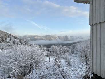 Árvores e arbustos cobertos de neve cobrem uma paisagem invernal. Montanhas e neblina surgem ao fundo sob um céu azul claro. Uma estrutura de madeira emoldura a cena.
