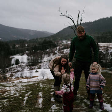 Uma família de quatro pessoas desfruta de um dia de inverno ao ar livre em uma colina gramada, cercada de neve, montanhas e árvores. O clima é alegre e descontraído.
