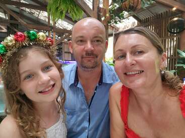 Eine lächelnde dreiköpfige Familie macht ein Selfie in einer festlichen Umgebung draußen. Das Mädchen hat ein Weihnachtsstirnband auf. Die fröhliche Szene ist von üppigem Grün umgeben.