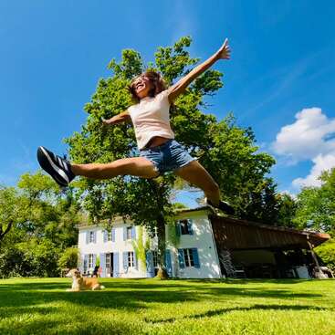A joyful woman jumps high in the air in a green yard, with a white house, blue shutters, trees, clear sky, and a relaxed dog.