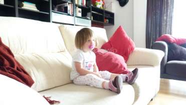 The image depicts a young girl sitting on a white couch, surrounded by red pillows and a bookshelf in the background.