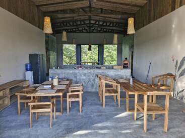 This open, airy kitchen features wooden furniture, pendant lights, concrete floors, and natural accents. Large windows offer a green view, with a person at the counter.