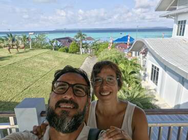A smiling couple takes a selfie on a sunny balcony, with lush gardens, palm trees, houses, and a stunning turquoise ocean in the scenic background.