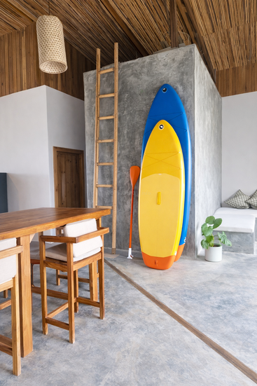 A modern room features a wooden dining table, cozy chairs, a plant, colorful surfboards, a paddle, a wooden ladder, and natural wooden accents throughout.