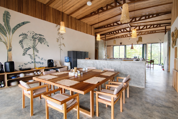 This image shows a modern, airy dining area with a large wooden table, cushioned chairs, natural lighting, hanging lamps, and tropical mural artwork on the wall.
