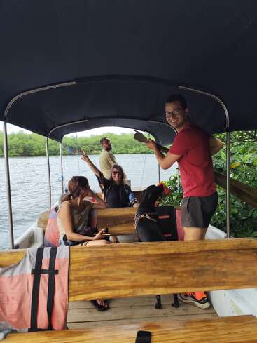 Four people and a dog are enjoying a boat ride surrounded by lush greenery. They are smiling, relaxing on wooden benches under a canopy, having fun.