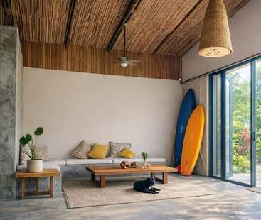 A cozy, modern living room with natural wood accents, surfboards, indoor plants, sliding glass doors, and a lounging black dog on a woven rug. Relaxing ambiance.