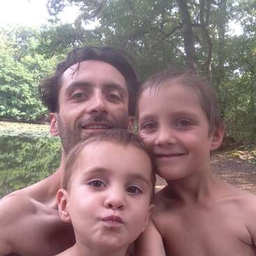 La imagen muestra a un hombre y dos niños posando para un selfie frente a un lago sereno, rodeados de frondosos árboles verdes en un día soleado.