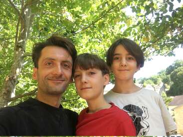 Ein Mann und zwei Jungen lächeln im Freien und stehen dicht beieinander unter grünen Bäumen. Das Sonnenlicht dringt durch und schafft eine helle und fröhliche Atmosphäre. Ein Familienmoment.