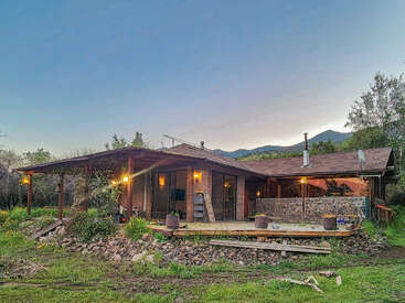A rustic wooden cabin with a wide porch, surrounded by greenery and mountains at sunset. Warm lights glow, creating a cozy, tranquil rural atmosphere outside.
