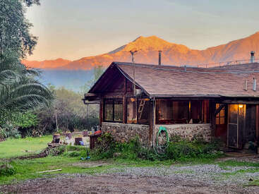 A rustic house with stone and wood exterior sits in a lush yard, with sunlit mountains in the background, creating a peaceful countryside setting at dusk.