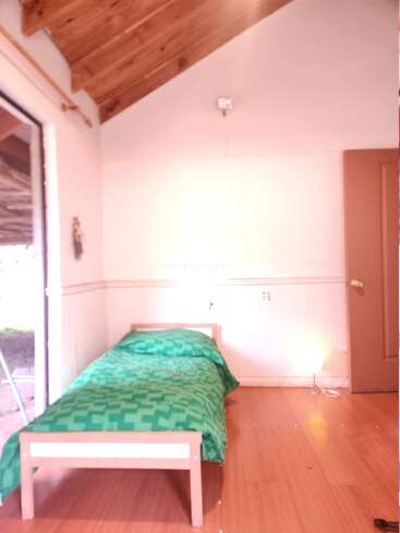 This image shows a simple, cozy room with a single bed covered by a green patterned blanket, wooden floor, slanted wooden ceiling, and an open door.