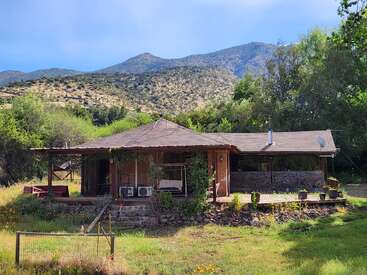 A rustic house with a wooden porch sits surrounded by vibrant greenery and wildflowers, nestled at the base of scenic, sunlit mountains under a clear blue sky.