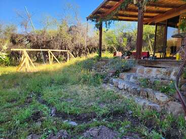 A peaceful backyard scene shows a rustic patio with chairs, stone steps, lush green grass, wildflowers, and a wooden swing frame under a clear blue sky.