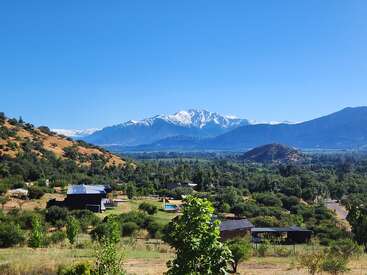 A lush green valley dotted with houses, trees, and hills under a clear blue sky. Snow-capped mountains rise majestically in the distant background. Peaceful scenery.