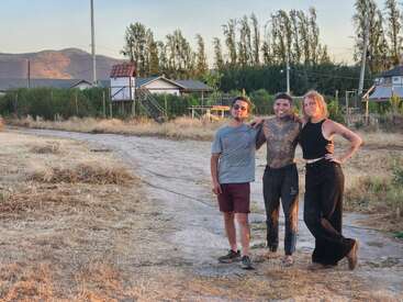 Three people pose together on a rural dirt path with dry grass, houses, and mountains in the background. They appear happy and relaxed in casual attire.