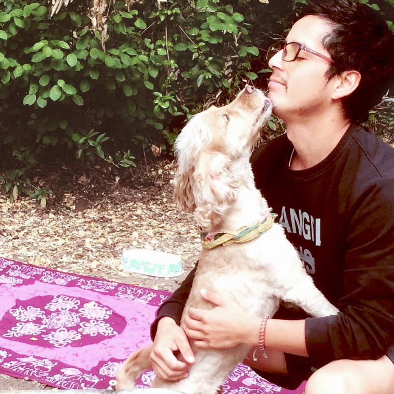 A man and his dog share a loving moment outdoors. The dog lovingly licks the man’s face while sitting on his lap, surrounded by greenery.