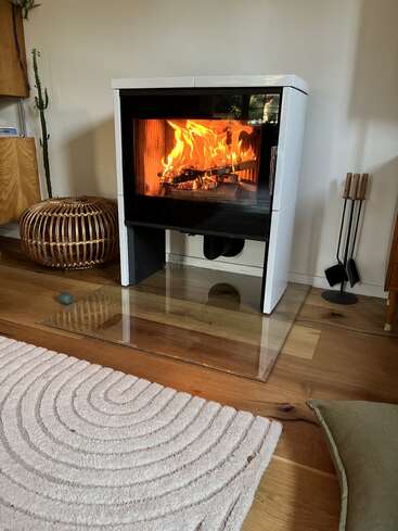 Ein moderner Holzofen mit einem hellen Feuer drinnen steht auf einem Glasherd in einem gemütlichen Wohnzimmer mit Holzböden, einem Teppich und Deko.