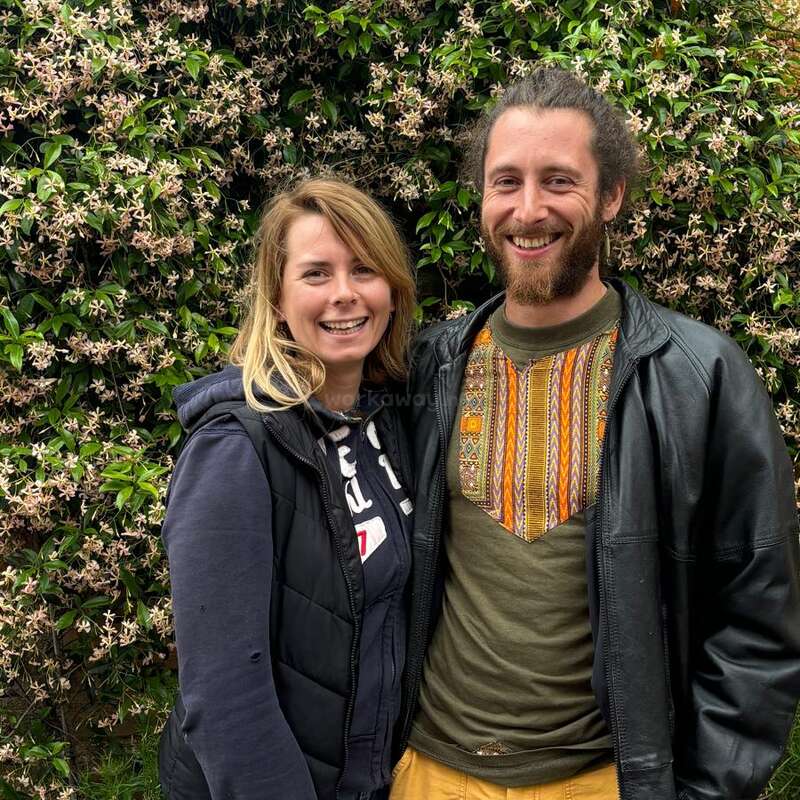 Ein lächelnder Mann und eine lächelnde Frau stehen eng beieinander im Freien vor einer üppigen, mit blühenden Blumen bewachsenen Wand. Sie sehen glücklich und entspannt aus und genießen die Natur.