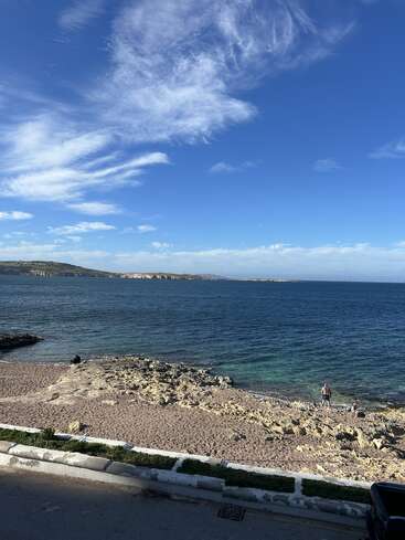 A peaceful coastal scene with clear blue sky, wispy clouds, calm sea, rocky shore, a sandy beach, and a few people enjoying the tranquil scenery.