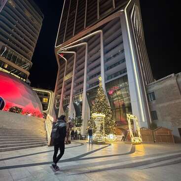 The image shows a modern city square at night with tall, uniquely designed buildings, a large decorated Christmas tree, festive lights, and people walking around peacefully.